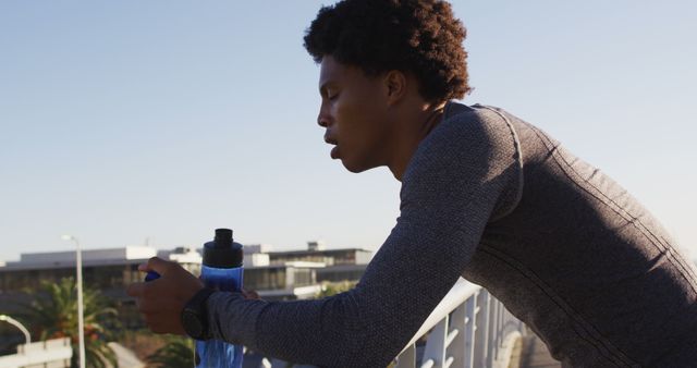 Fit african american man exercising in city, resting on footbridge, drinking from water bottle. fitness and active urban outdoor lifestyle.