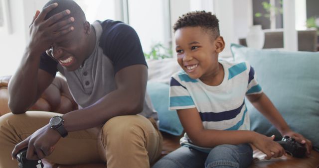 A father and his son are playing video games on a sofa in their living room. The father shows a defeated expression while the son is smiling joyfully. This scene depicts quality family time, bonding, and the fun of video gaming. Perfect for use in family lifestyle promotions, gaming advertisements, or articles about parenting and technology.