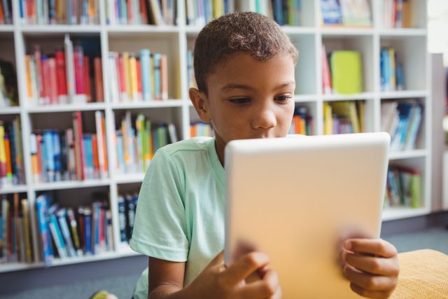 Young boy using a tablet in a library, surrounded by bookshelves. Ideal for educational content, technology in education, digital learning, and modern classroom settings.