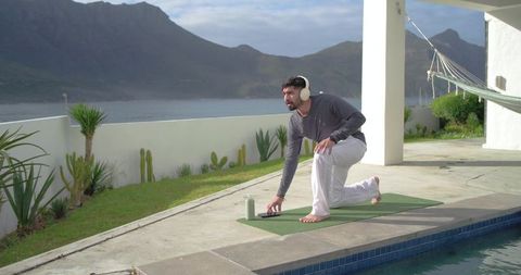 Indian man practicing yoga on coastal pool terrace wearing wireless headphones