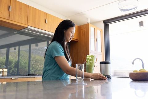 Asian woman preparing green smoothie at sunlit kitchen island for wellness lifestyle