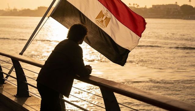 Silhouette man leaning on promenade with egyptian flag at sunset, backlit reflections