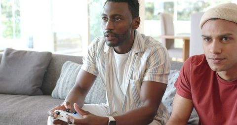Diverse friends playing console game on living room sofa, focused Black and Latino men