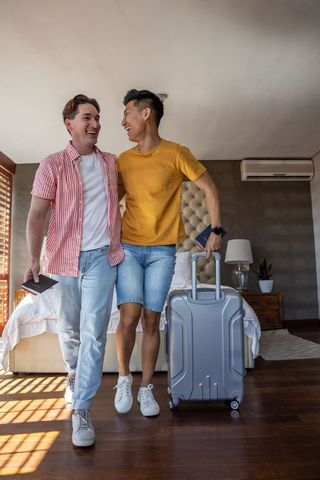 Smiling gay couple walking in sunlit hotel room holding passports and silver suitcase