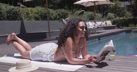 African american woman reading and sunbathing poolside on sunlit deck with straw hat