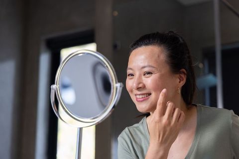 Mature asian woman examining skin in magnifying mirror, smiling, skincare routine