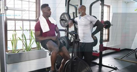 Man pedaling on air bike while coach leaning on squat rack and giving workout tips