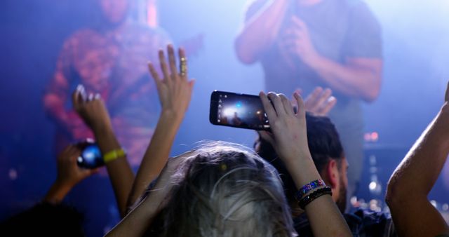 Concertgoers capture the live performance on their smartphones, with the stage lights creating a vibrant backdrop. The energy and excitement of a live music event are palpable as fans record memories.