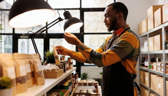 Retail owner adjusting lamp while inspecting artisan packaged goods on shelves