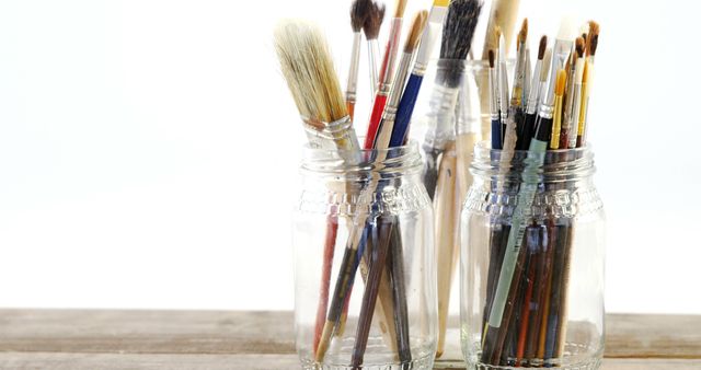 This image features a collection of varied paintbrushes stored in glass jars on a wooden table. Perfect for art and craft-related content, blog posts about painting techniques, or advertisements for creative materials.