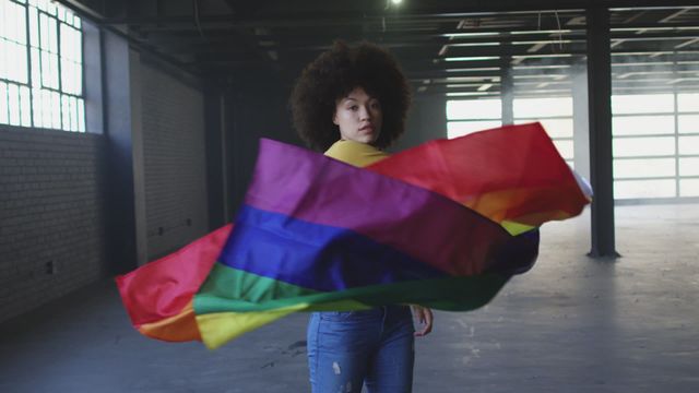 Gender fluid person with afro hairstyle holding rainbow flag in spacious, empty building. Colorful flag symbolizing LGBTQ pride and diversity. Great for use in campaigns about equality, LGBTQ rights, empowerment, and inclusion.