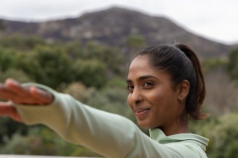 Smiling indian woman stretching arm on terrace wearing green top, ponytail, nose stud, outdoors
