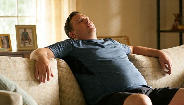 Middle-aged man dozing on beige sofa in blue tee and shorts relaxed afternoon nap