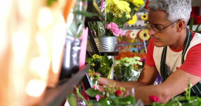 Senior male florist arranging flowers on a display shelf in a flower shop. Bright, colorful flowers in various stages of arrangement surround him. Useful for concepts like small business, gardening, retail, flower arrangement, and senior employment.