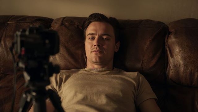 Reclining young man recording self facing tripod camera on leather couch in warm interior