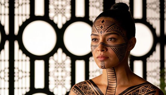 Woman posing with geometric tribal body paint and bold facial designs, backlit screen