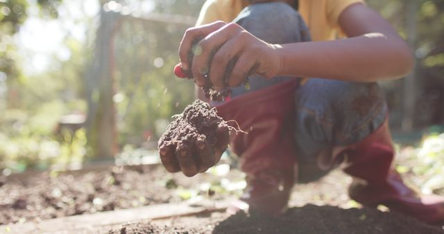 Boy kneeling in garden carefully holding soil with hand, embodying themes of gardening, agriculture, sustainability. Sunlight filters through trees, creating a serene and warm atmosphere. Ideal for promoting gardening, environmental awareness, children’s outdoor activities, educational content on sustainability and nature.