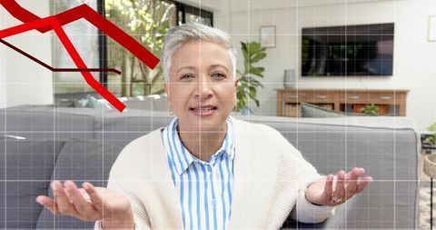 Senior woman explaining finances on home sofa, gesturing with falling red graph overlay