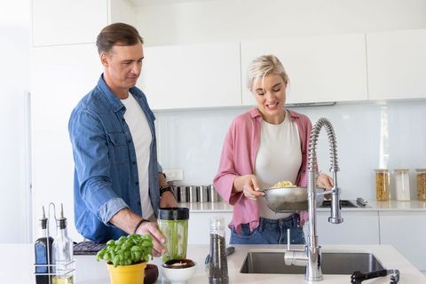 Couple cooking together in modern kitchen draining pasta and blending smoothie