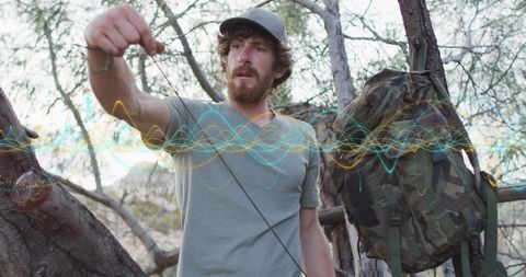 Bearded hiker securing survival cord across tree trunks with camo backpack in scrubland