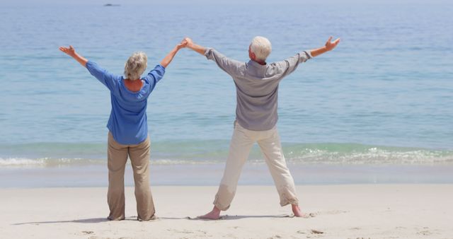 The image depicts an elderly couple standing on the beach with their backs to the camera and arms raised in a joyful gesture. They appear to be enjoying a peaceful and sunny day by the sea, with clear blue waters and white sand. Ideal for cases where themes of retirement, happiness, togetherness, and the beauty of nature are needed. Perfect for use in travel promotions, retirement living ads, health and wellness content, or inspirational materials.