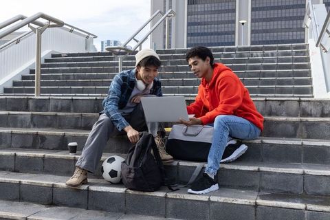 Asian and black teen male students collaborating on laptop on urban steps with soccer ball