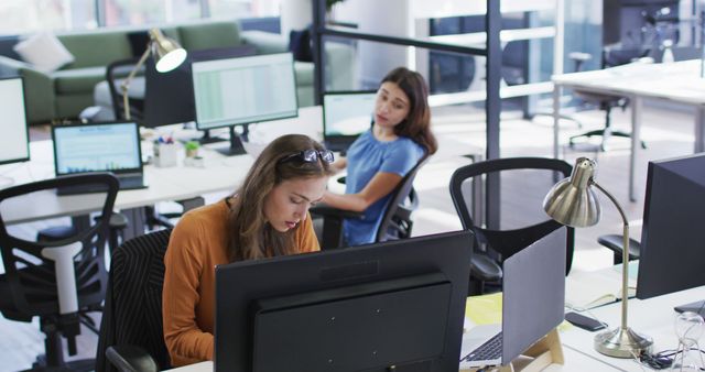 Two caucasian businesswomen sitting at desks using computers turning around to talk to each other. independent creative business in a modern office.