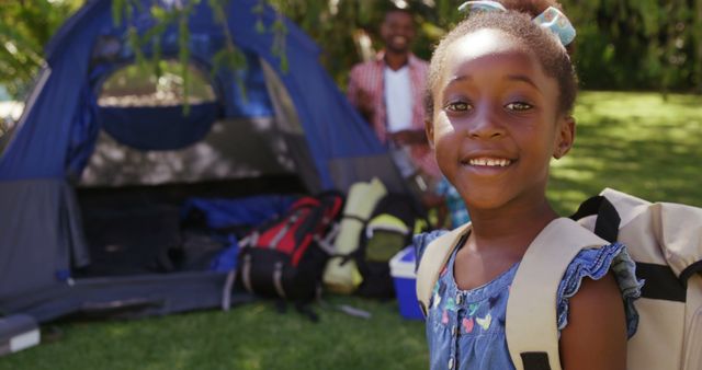 Young African American girl smiling near a tent with a backpack, suggesting she is on a camping trip with family. The background features outdoor camping gear and an adult, indicating a summer vacation setting. Ideal for use in advertisements for family vacations, camping gear, or articles about outdoor activities and bonding.