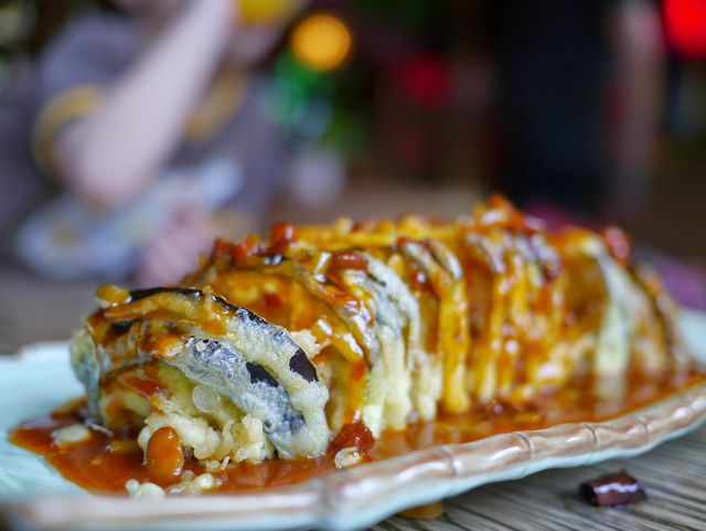This image captures a close-up view of a beautifully presented tempura sushi roll generously covered with sauce. Perfect for use in articles or advertisements about Japanese cuisine, gourmet food presentations, food blogs, or restaurant promotions. It highlights the flavors and quality of traditional Asian dishes and can be appealing for food enthusiasts and culinary publications.