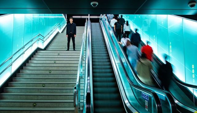 Solo commuter standing on stairwell beside cyan-lit escalator in underground transit hub