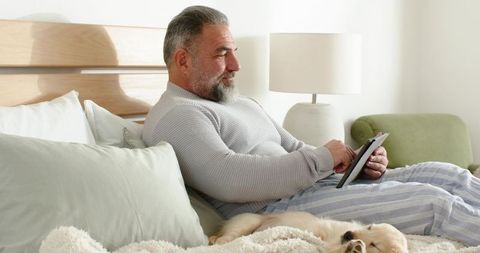 Senior man relaxing in bed using tablet with sleeping dog, cozy morning routine