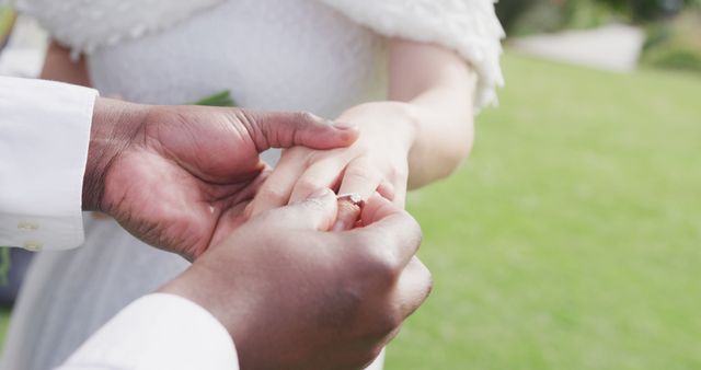 Image of hands of diverse bride and groom, groom putting ring on bride's finger at outdoor wedding. Marriage, love, happiness and inclusivity concept.