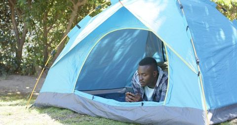 African american man relaxing in blue tent using smartphone during sunny forest camping