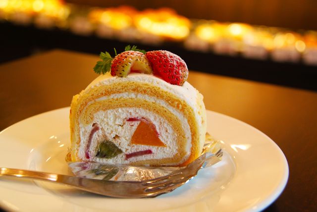 Close-up of a slice of strawberry Swiss roll cake on a white plate with a fork. The cake features fresh fruit slices and creamy filling. Perfect for food blogs, dessert menus, bakeries, and social media posts related to sweets and confections.