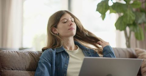 Young woman relaxing on sofa with laptop pushing hair back working remote sunlit