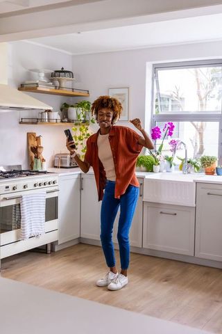 African american woman dancing in bright modern kitchen holding smartphone enjoying music