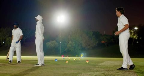 Night cricket under floodlights: bowler running, batsman with navy helmet and floating emojis