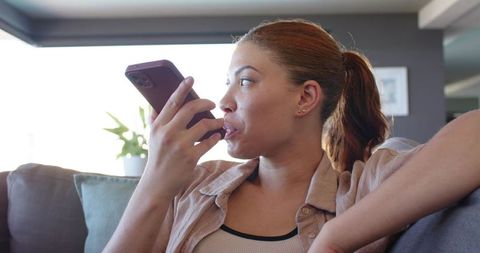 Woman speaking into smartphone while relaxing on sofa, recording voice message at home