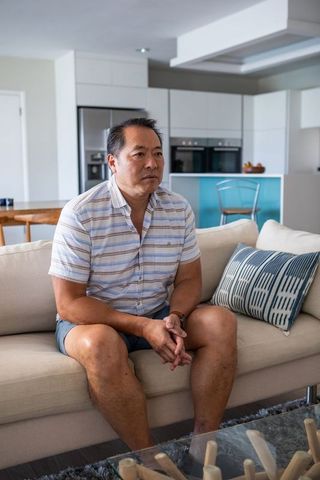 Middle-Aged Asian Man Relaxing on Modern Beige Sofa in Bright Living Room