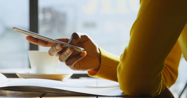 Person using a digital tablet while sitting in a cozy café. Hand in a yellow sweater browsing or reading on the touchscreen. Ideal for use in technology, lifestyle articles, café ambiance promotions, and remote working concepts.