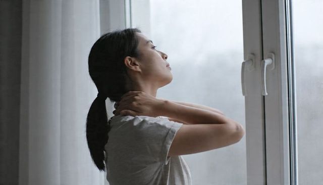 Adult asian woman massaging neck while gazing out window in quiet apartment