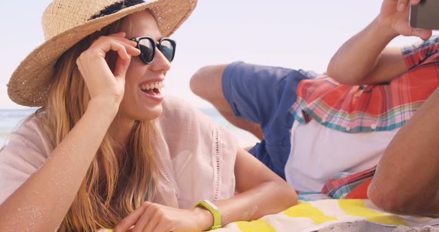 Young woman relaxing on beach towel wearing sunglasses and hat, smiling at friend. Ideal for promoting summer vacations, travel destinations, beachwear, and lifestyle products. Use in marketing campaigns related to leisure and holidays.