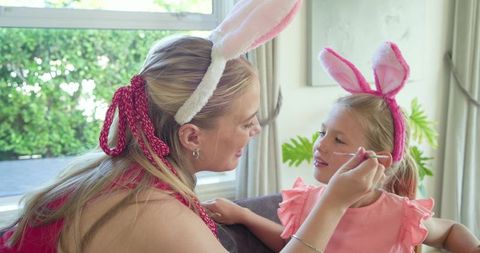 Mother painting daughter face wearing bunny ear headbands for easter family bonding