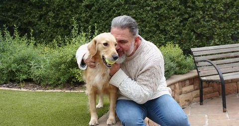 Senior man cuddling golden retriever holding tennis ball on backyard lawn near bench