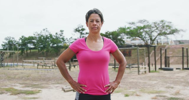 Confident woman standing in an outdoor fitness park, hands on hips, wearing a pink shirt. Ideal for promoting outdoor fitness activities, health and wellness programs, active lifestyle campaigns, and exercise motivation.