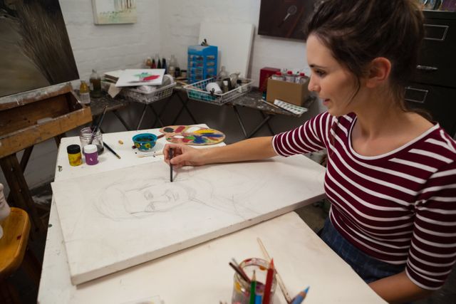 Young woman attentively sketching on a canvas in an art class. She is surrounded by various art supplies and appears focused on her work. This image can be used for promoting art classes, workshops, creative hobbies, or educational materials related to art and drawing.