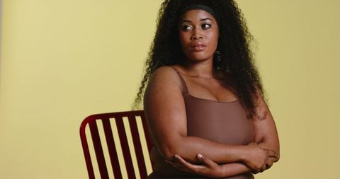 Contemplative african american woman sitting on red chair wearing brown dress and headband