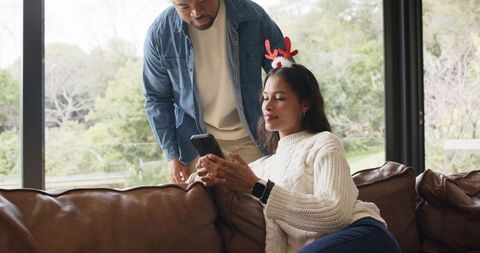 Couple relaxing on leather sofa while woman checking smartphone wearing reindeer antlers