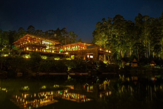 Nighttime view of luxury resort beautifully illuminated near a forest lake. Modern architecture with extensive use of glass, cozy lighting reflecting on calm water, creating a tranquil and inviting environment. Suitable for marketing material for travel agencies, promotional use for resorts, holiday advertisements, and nature retreats.