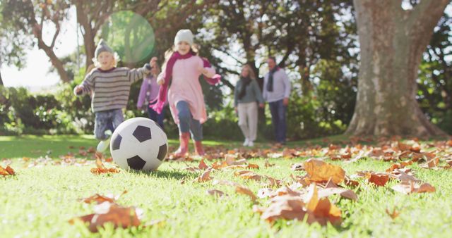 Children having fun playing soccer in a park surrounded by fallen autumn leaves. A tiny family is calmly strolling in the background. Great for depicting carefree childhood moments, sibling relationships, family outings, and outdoor activities in nature during the fall season.